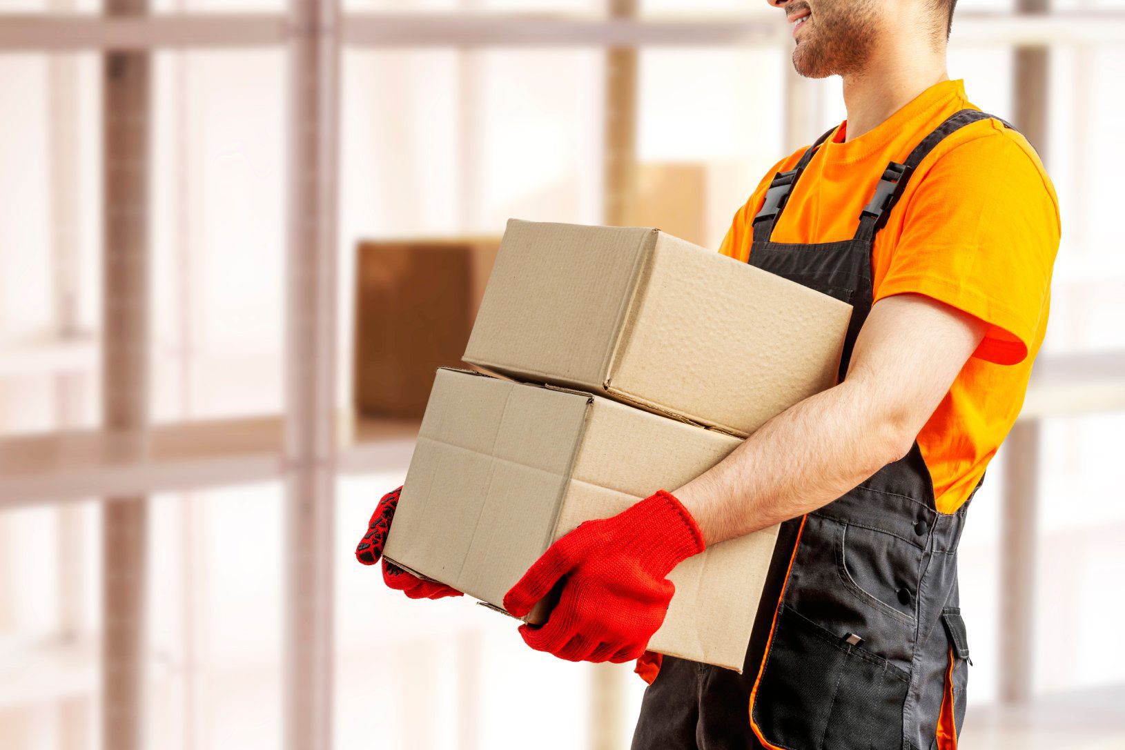 Young man holding cardboard package working in warehouse among racks and shelves. Delivery man with box. Staff laborer, orange uniform cap, t-shirt, coveralls service moving delivering orders goods.