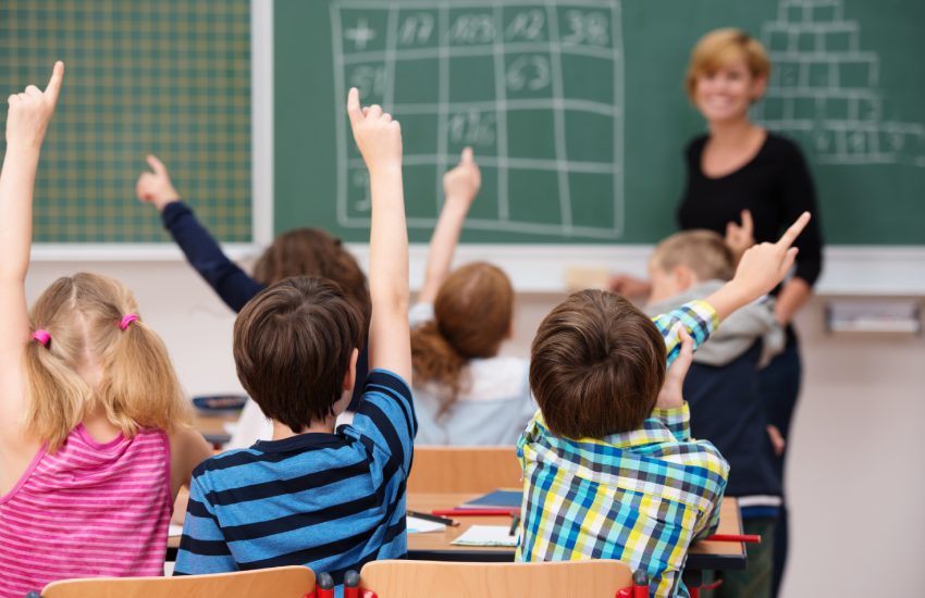 Intelligent group of young school children all raising their hands in the air to answer a question posed by the female teacher, view from behind