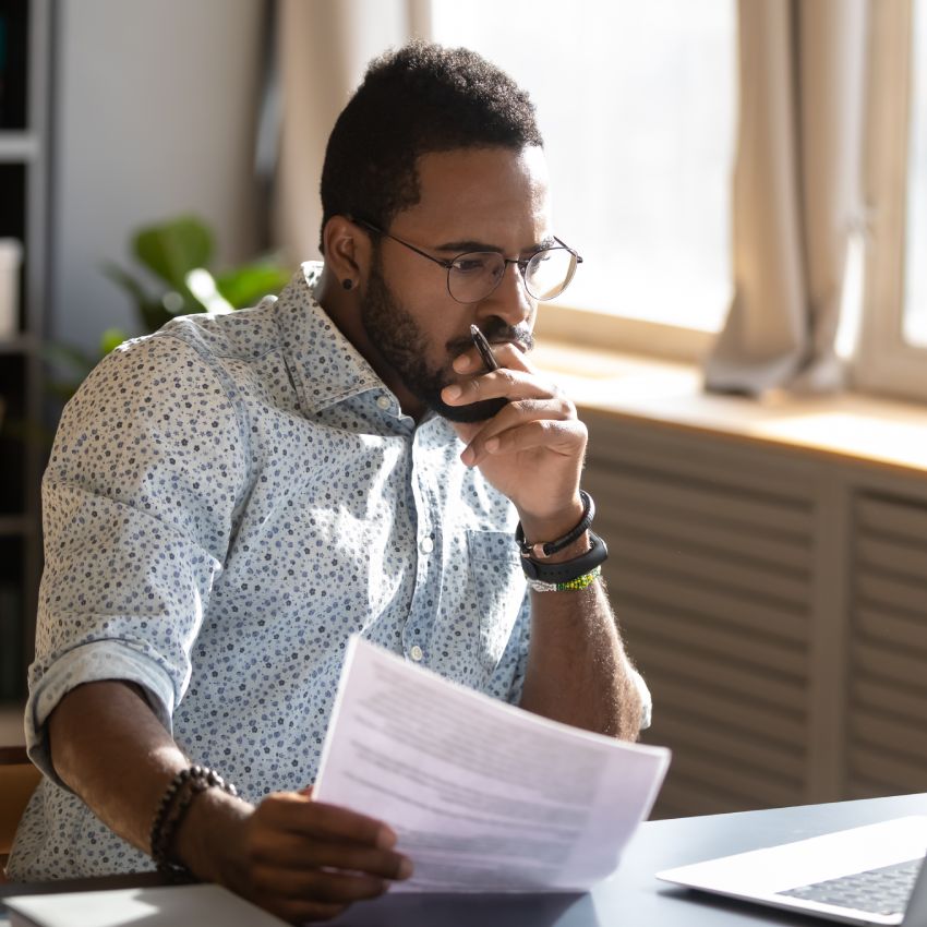 Focused serious african american businessman accountant analyst holding documents looking at laptop computer screen doing online trade market tech research thinking working sit at home office desk