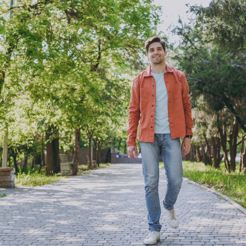 Full length young man 20s he wearing orange jacket blue t-shirt walking look camera rest relax in spring green city park go down alley sunshine lawn outdoors on nature. Urban lifestyle leisure concept