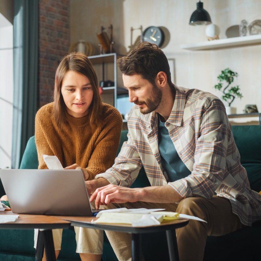 Stressful Accounting at Home: Couple Using Laptop Computer, Sitting on Sofa in Apartment. Young Family Filling Tax Forms, Mortgage Documents, Bills, Checks, Balances, Invoices are in Order