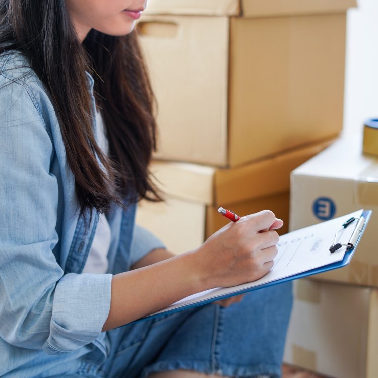 Asian woman checking and packing the carton box prepare to move to new house relocation shipping or check goods package before delivery to customer from online shopping by checklist