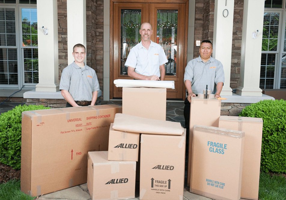 A group of men standing next to a pile of Allied moving boxes in front of an entry house