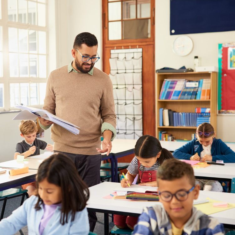 Primary school teacher walking in classroom while multiethnic students studying. Male teacher holding textbook helping schoolchildren during class. Professional educator walking in classroom.