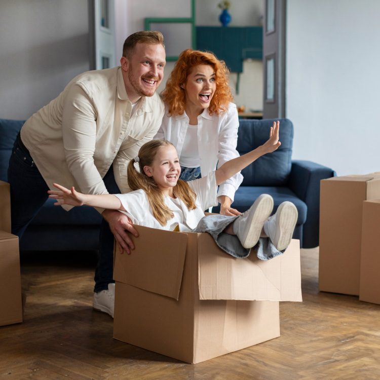 Happy family of three having fun and celebrating moving day, father and mother riding smiling daughter in cardboard box, fooling around in new flat