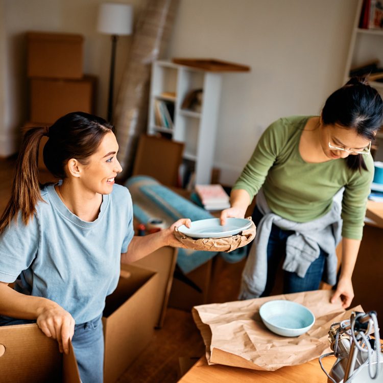 Happy woman and her Asian female friend wrapping fragile things into a paper while preparing to move out of the apartment.