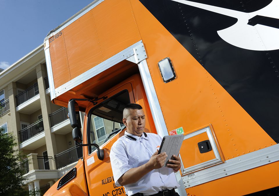 A man stands in front of a moving truck, holding a tablet and looking at the screen with a focused expression