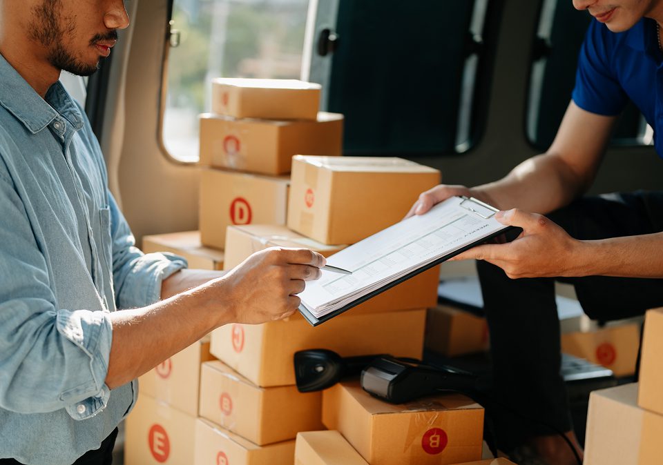 Two men are checking a moving dispatch checklist on a clipboard and carrying boxes in a logistics environment