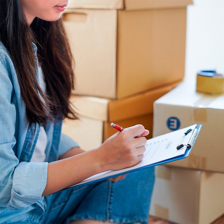A woman sits on the floor surrounded by boxes, holding a clipboard, preparing for a summer move in Minnesota.