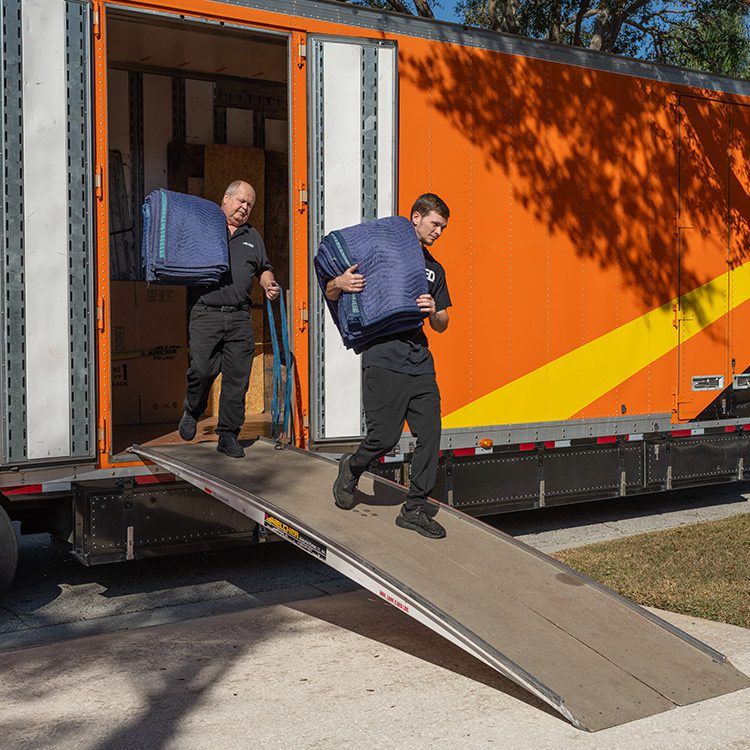 Two men load a large suitcase into a moving truck under the summer sun, preparing for a move in Minnesota.