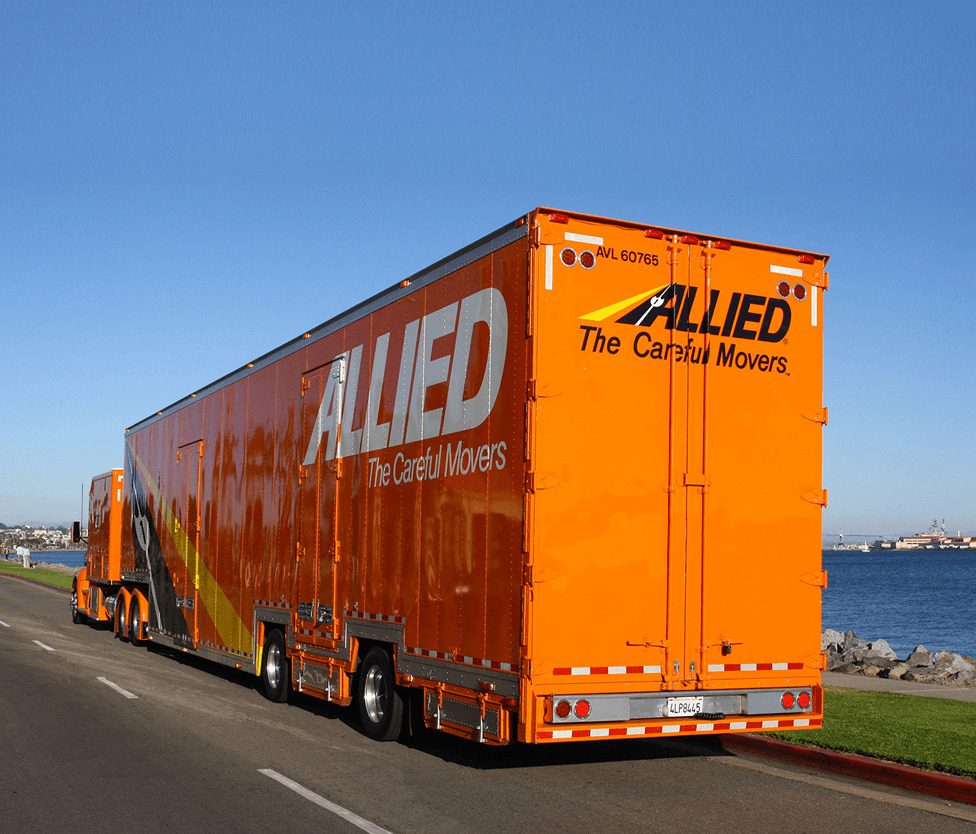 An orange Allied moving truck parked along a waterfront road, with a clear blue sky in the background.