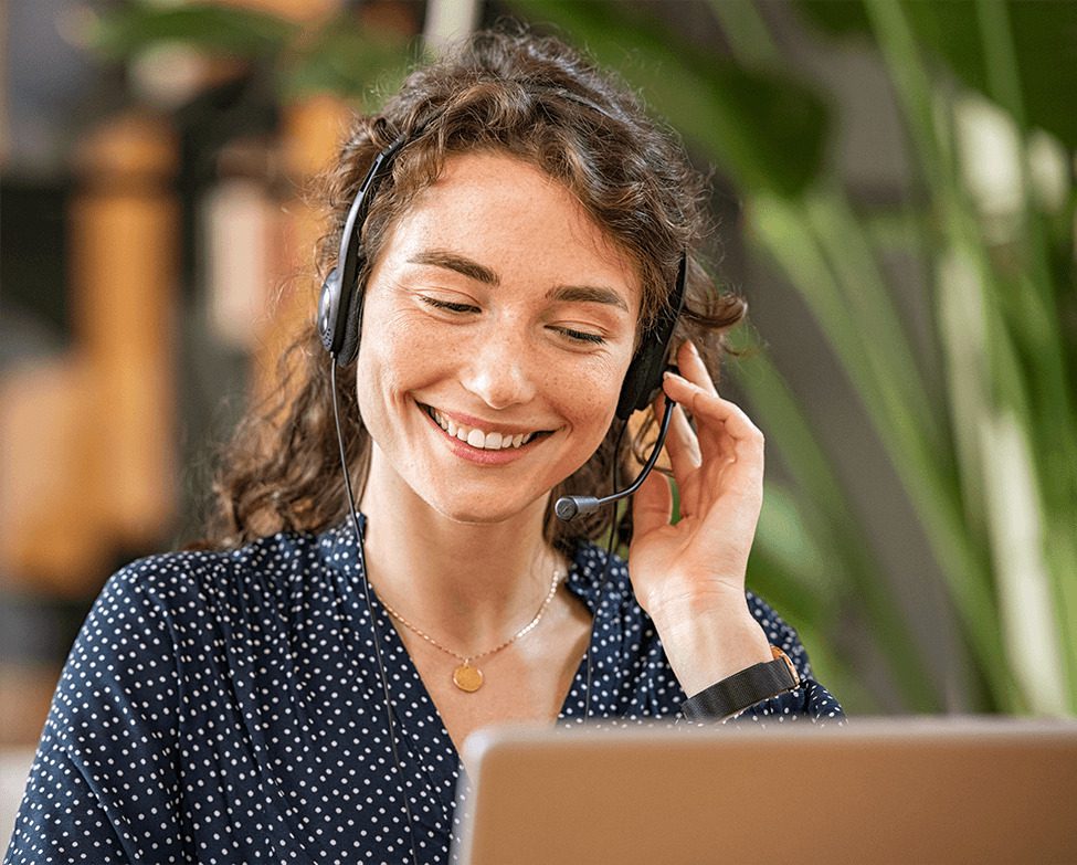 A woman with curly hair wearing a headset smiles while looking at a laptop in a bright, modern workspace.
