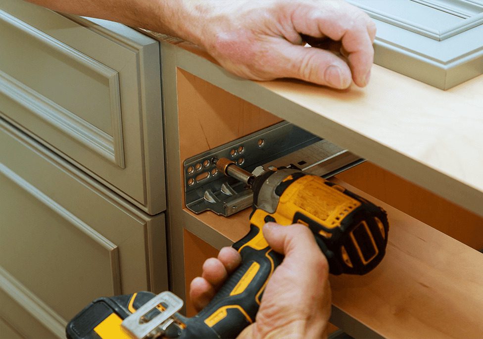 A person using a power drill to install a drawer slide on a wooden cabinet.