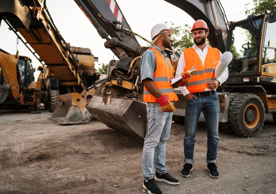 Two construction workers in safety vests and helmets discussing plans near heavy machinery on a job site.