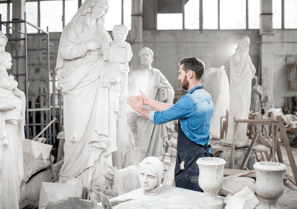 A sculptor in a workshop shapes a statue, surrounded by various stone sculptures and tools, with dust in the air.