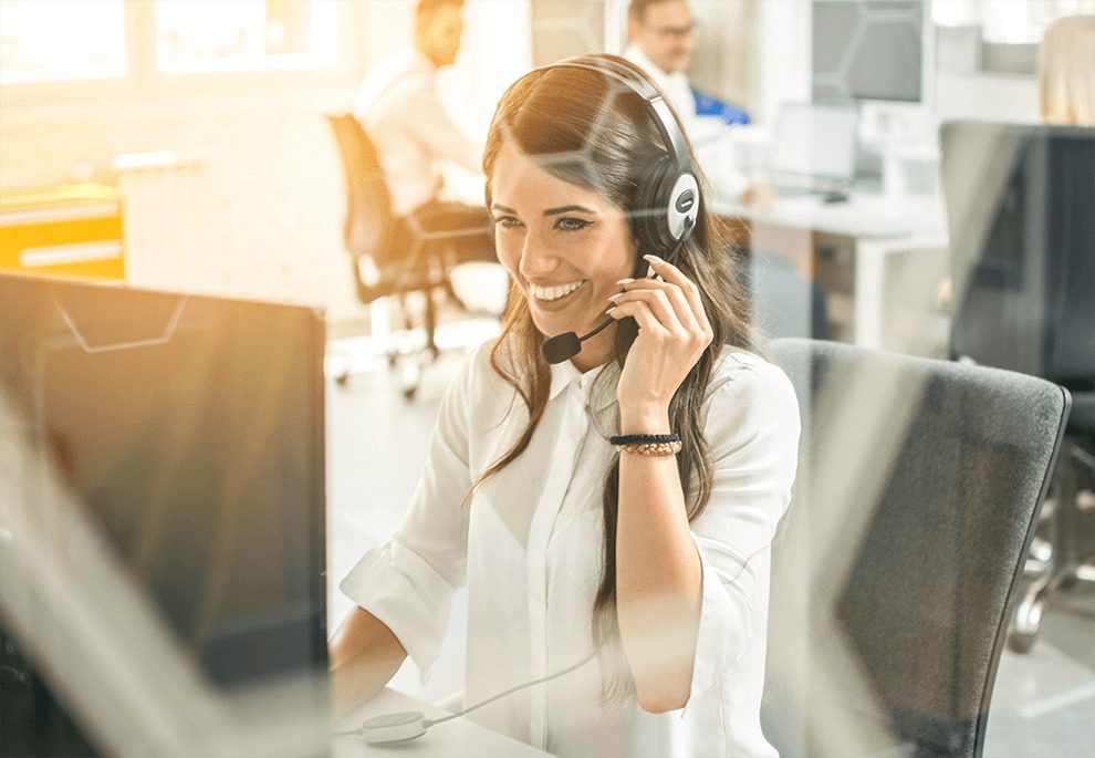 A woman wearing a headset smiles while working at a computer in a modern office environment.