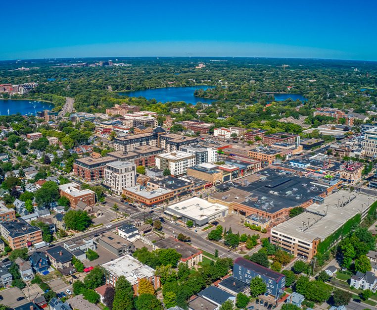 Aerial view of a vibrant urban area with mixed-use buildings, green parks, and a lake in the background under a clear blue sky.