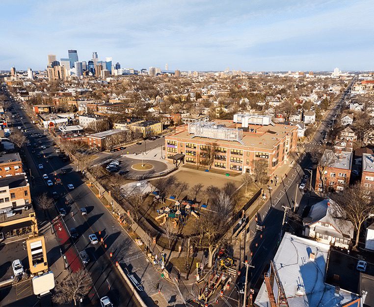 Aerial view of a city neighborhood featuring a school, playground, and surrounding residential areas with a skyline in the background.
