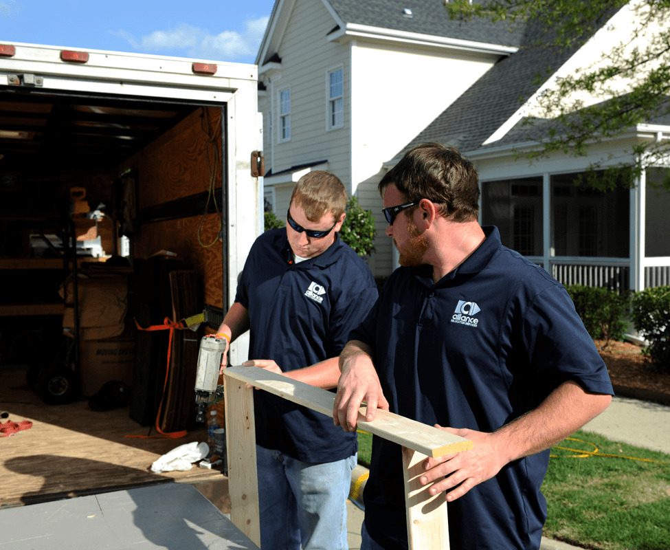 Two movers in navy blue shirts work together on a wooden frame outside a house, with a truck and tools in the background.