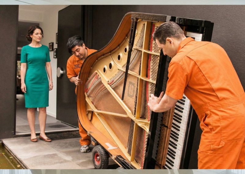 Two piano movers carefully moving a grand piano while a woman watches.