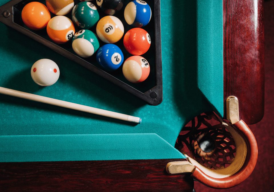 A close-up view of a pool table with a triangle rack of colorful billiard balls and a cue stick resting on the green felt.