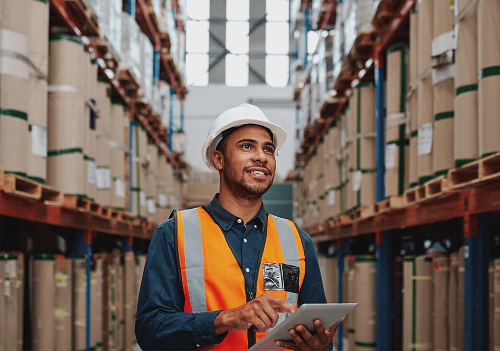 A warehouse worker in a hard hat and safety vest uses a tablet while standing between rows of stacked boxes.