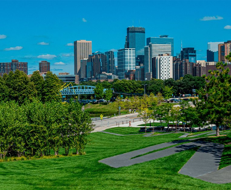 City skyline with modern buildings under a clear blue sky, surrounded by green parks and trees.