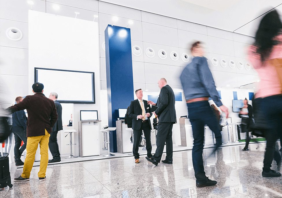 Busy conference hall with professionals networking, standing near booths and screens, creating a dynamic atmosphere.