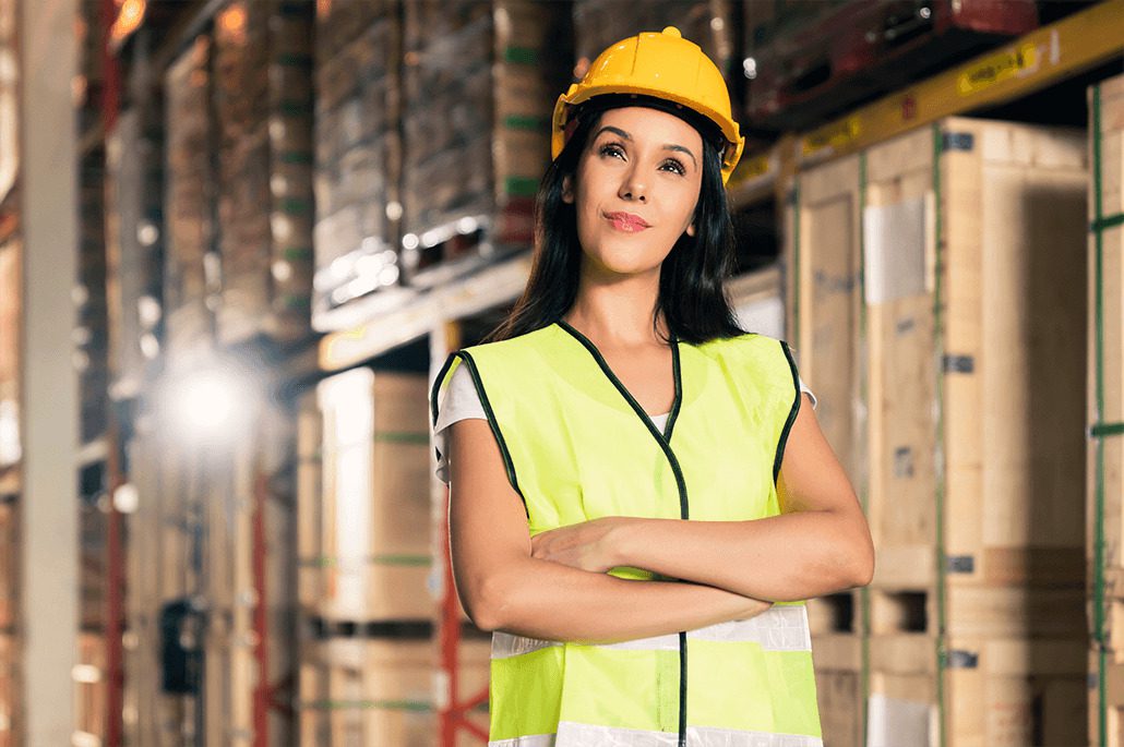 A confident woman in a yellow hard hat and reflective safety vest stands with arms crossed in a warehouse filled with stacked pallets.