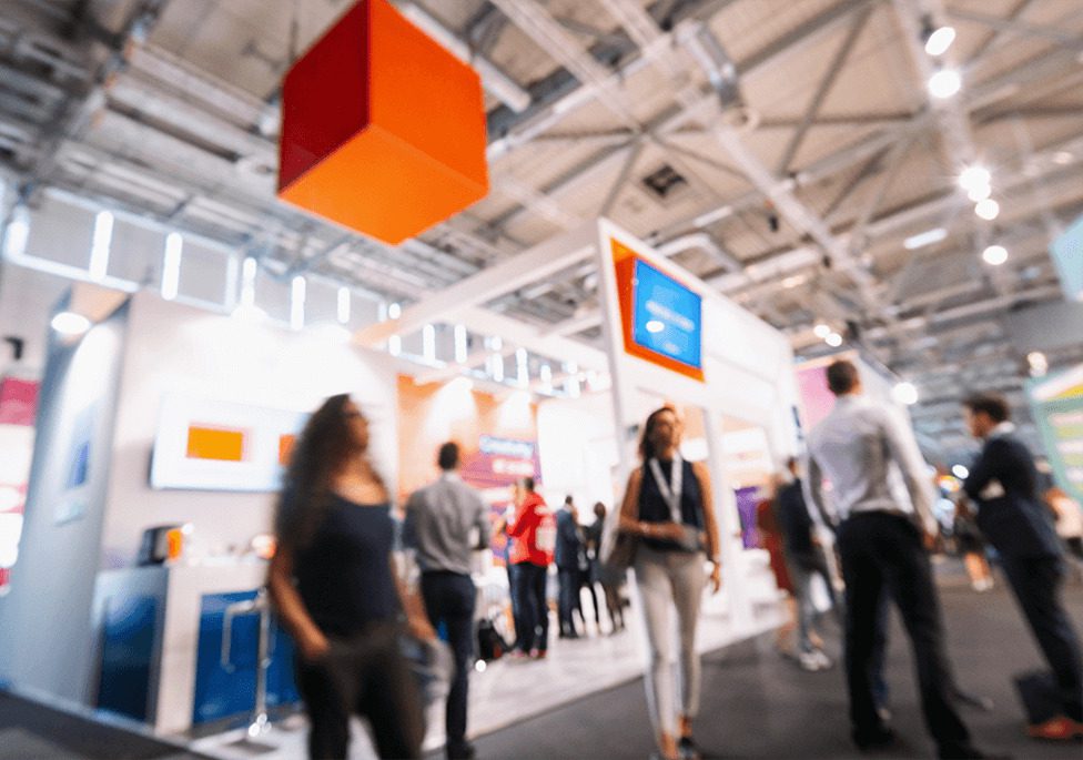 A busy exhibition hall with blurred figures walking past colorful booths and displays.