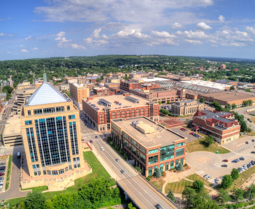 Aerial view of a cityscape featuring modern buildings, roads, and green spaces under a partly cloudy sky.