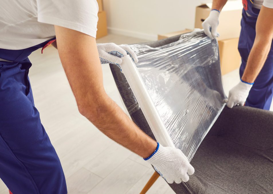 Skilled movers are captured in a close up shot, wrapping an armchair in protective stretch film during home or office moving. This attention to securing of furniture ensures a damage free service.