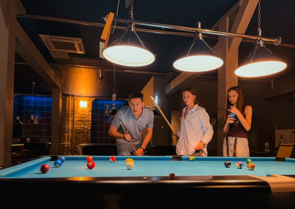 Father playing billiards with two teenage daughters. Family spending leisure time together, enjoying indoor sport, fun, teamwork, and communication. Concept of family recreation, parenting.
