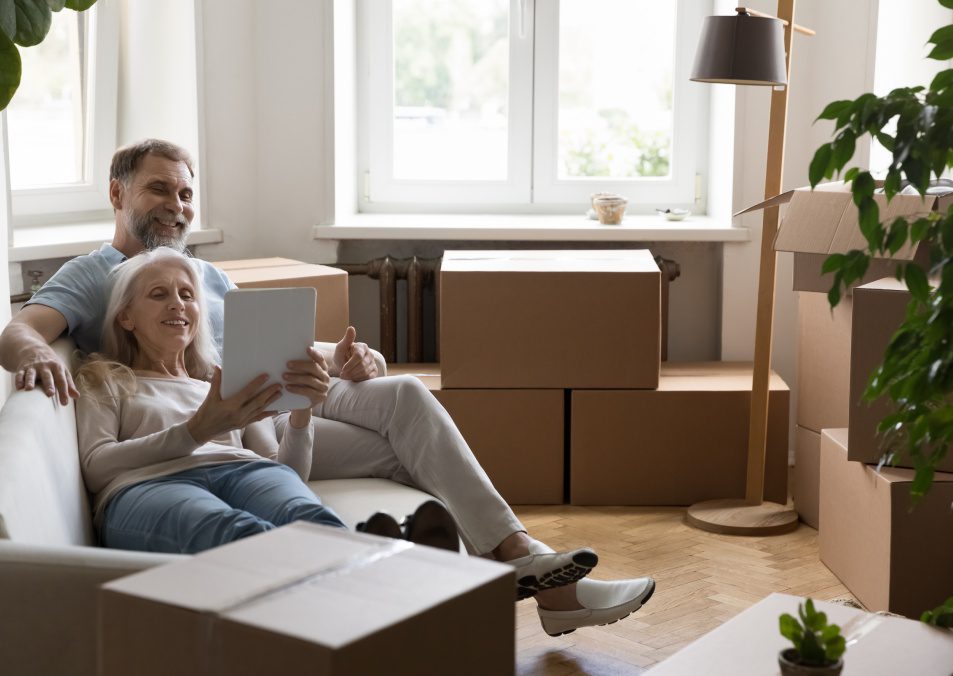 Mature couple using digital tablet buying goods for new home relaxing on cozy couch in living room with packed cardboard boxes with stuff on relocation day. Do easy comfort remote e-shopping concept