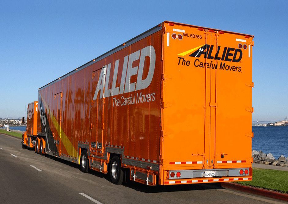 An orange Allied moving truck parked along a waterfront road, with a clear blue sky in the background.